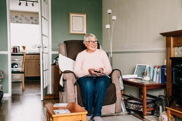 A smiling senior woman changes channels on the remote control while watching TV at home