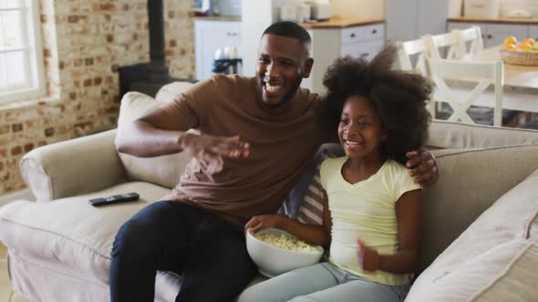 African american daughter and her father on couch eating popcorn watching tv and high fiving alt