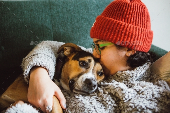 Young woman in an orange beanie and comfy sweater snuggling a comfy dog on the couch - Stock Photo - Images