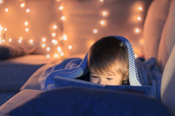 Cute little baby boy reading a book lying on sofa under knitted blanket with garland lights at