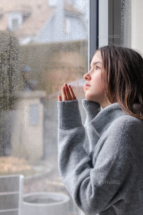 Tween thoughtful girl in gray sweater looking through the window at rainy sky with hope