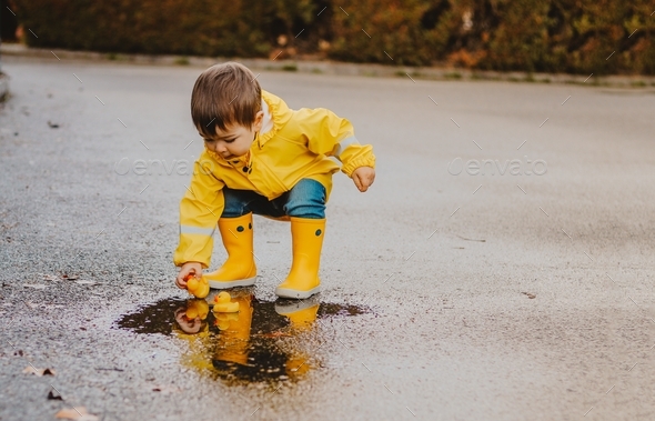 Little boy in yellow rubber boots playing with duck toys in the puddle after rain