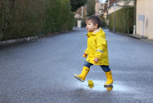 Cute playful little baby boy in bright yellow raincoat and rubber boots playing with toy boat and
