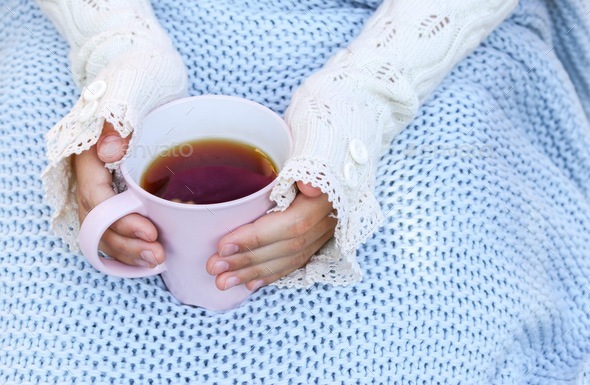 Hands of little girl in cozy hand warmers fingerless gloves holding cup of tea on her knees wrapped