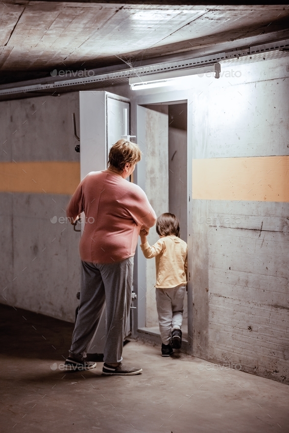 Senior woman with little child entering nuclear fallout shelter built under the house