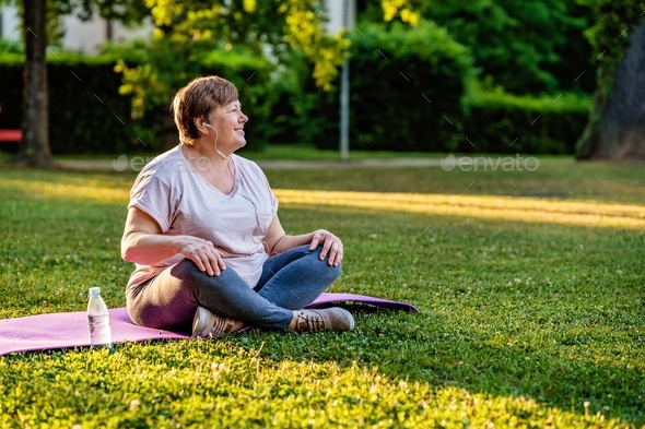 Relaxed senior plus size woman with earphones sitting on yoga mat outdoors  resting after exercises Stock Photo by Kiwitanya