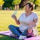 Relaxed senior plus size woman with earphones sitting on yoga mat outdoors  resting after exercises Stock Photo by Kiwitanya