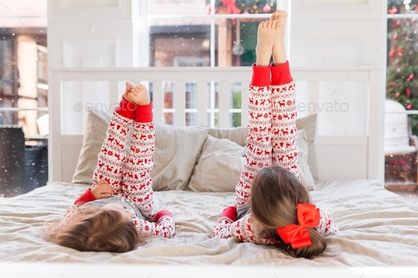Two girls in xmas pajamas lying on the bed and lifting their legs up. Christmas moments with kids