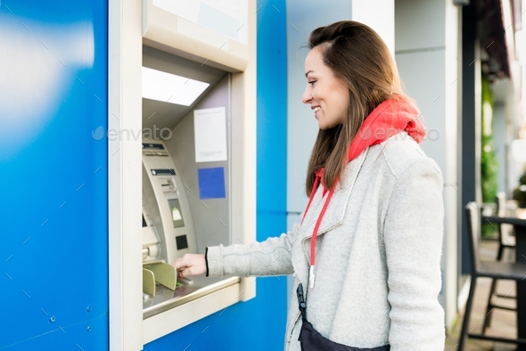 Young brunette woman inserting credit card, withdrawing money at outdoor ATM.