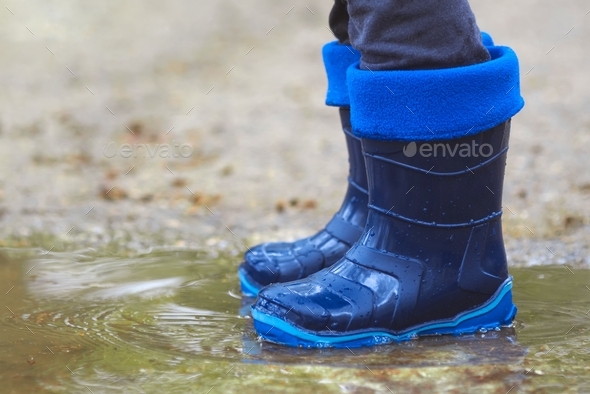 A child boy in a blue waterproof rubber boots runs through the puddles in the city