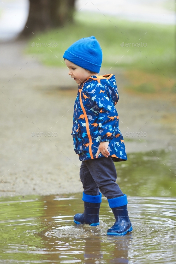 A child boy in a waterproof jacket and rubber boots runs through the puddles in the city