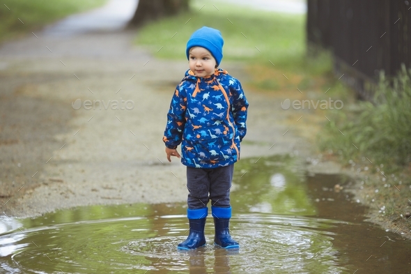 A child boy in a waterproof jacket and rubber boots runs through the puddles in the city