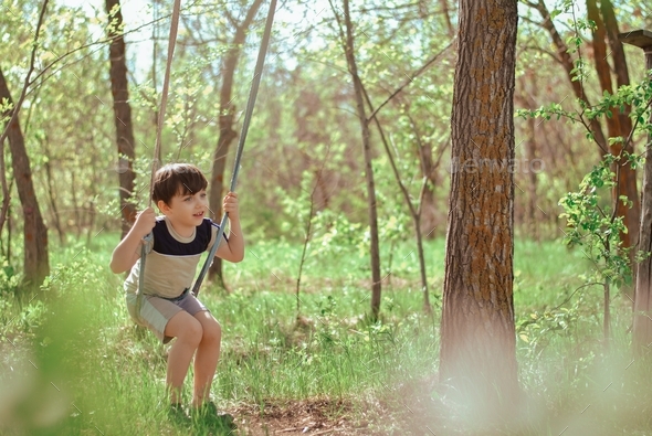 Happy smiling kid little boy rides on a rope swing in the spring forest. Happiness of childhood