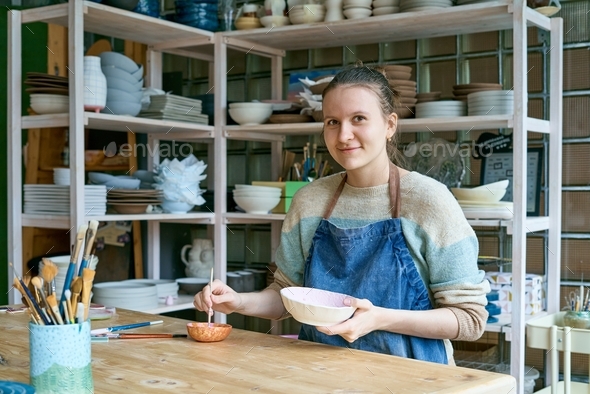 Skilled young woman in apron standing at table and drawing on ceramic bowl in pottery workshop