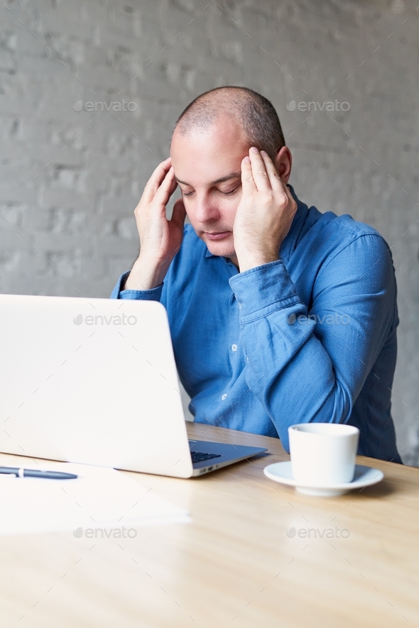 A man suffers a headache, sits at work in the office at a laptop, rubs his hands with whiskey
