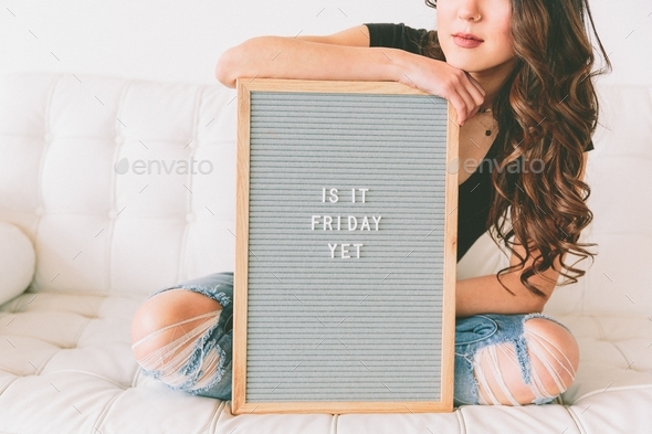 A woman sitting on a couch while holding a sign that says \'is it Friday yet\'.