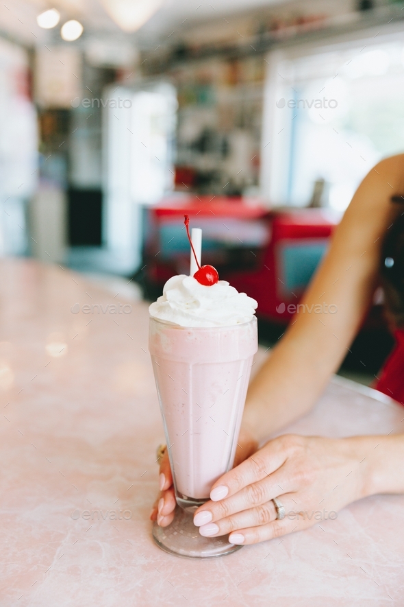 A woman holding a pink strawberry milkshake on a diner counter.