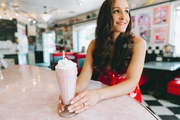A beautiful young woman with a strawberry milkshake sitting at a counter in a retro diner.