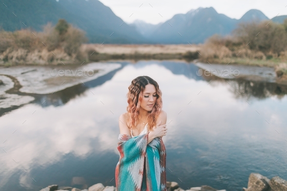 A beautiful woman with pink hair wrapped in a blanket standing in front of a mountain and lake. - Stock Photo - Images