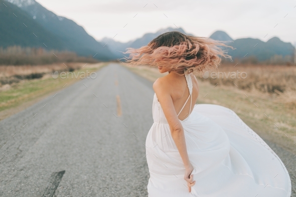 A woman with pink hair in a white dress twirling on a street in the mountains. - Stock Photo - Images