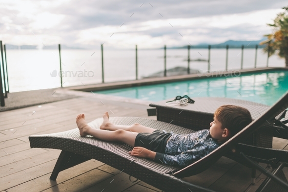 A little boy lounging and relaxing on a deck chair by a swimming pool by the ocean.  - Stock Photo - Images