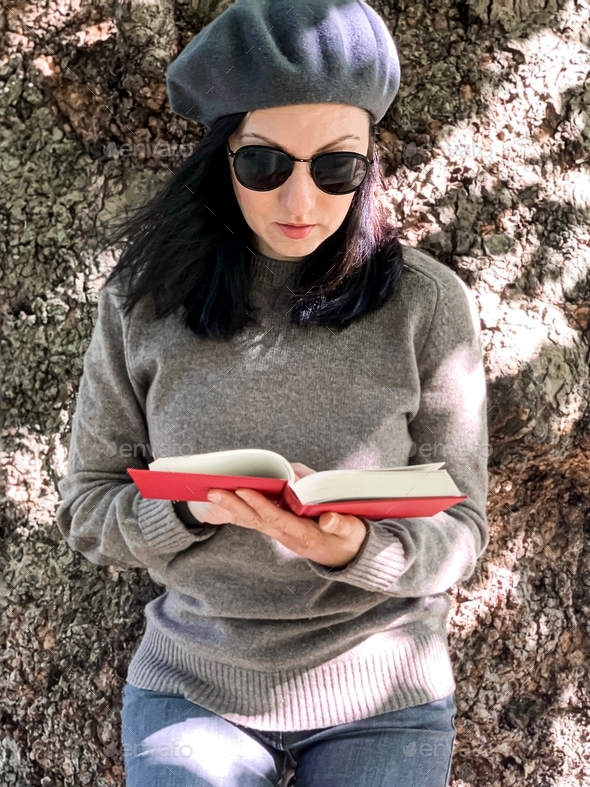 Young Millennial Candid Woman In French Beret Is Reading Red Book Standing By Huge Tree In A 