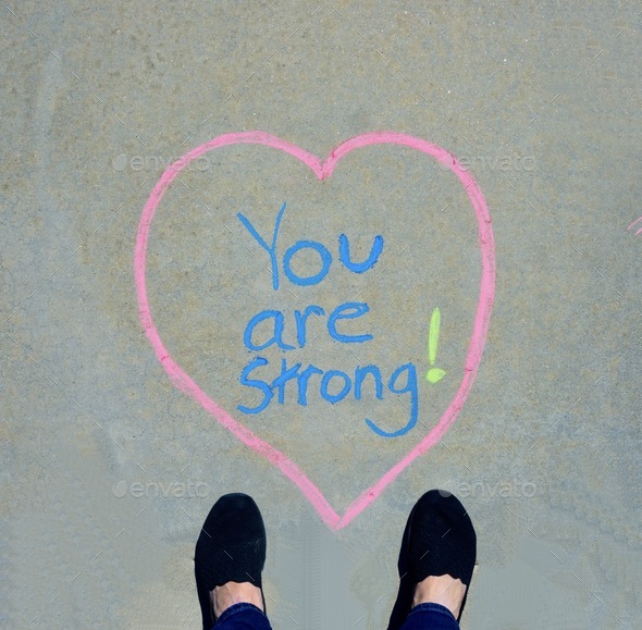 Looking down at a sidewalk with “You are strong” written in blue in the middle and black shoes