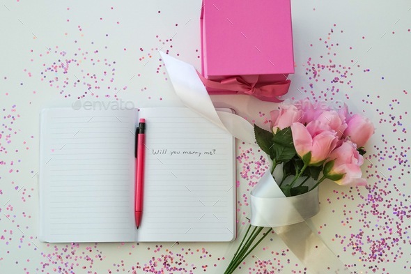 Open journal with proposal , pink pencil, pink gift box and pink flowers.