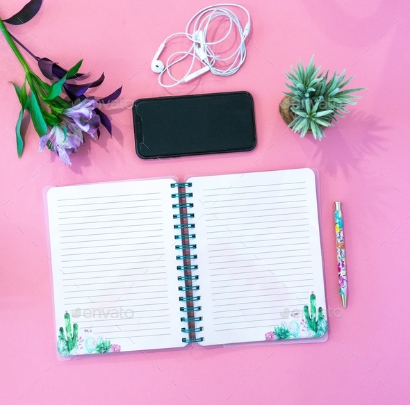 Overhead view of an open blank journal, pen, phone, earbuds, plant and flowers on a pink background