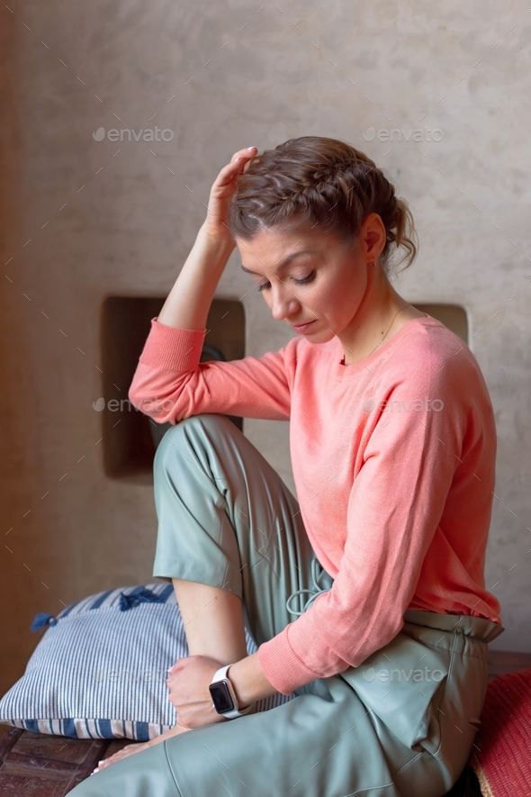 pensive woman in coral sweatshirt and gray pants is sitting in the room