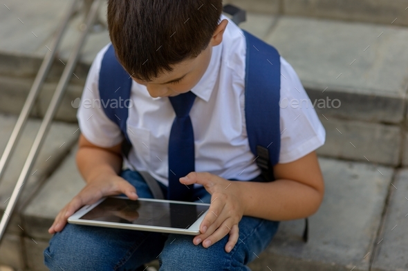 A brunet schoolboy with a knapsack behind his back sits on the stairs and plays a tablet. Close up - Stock Photo - Images
