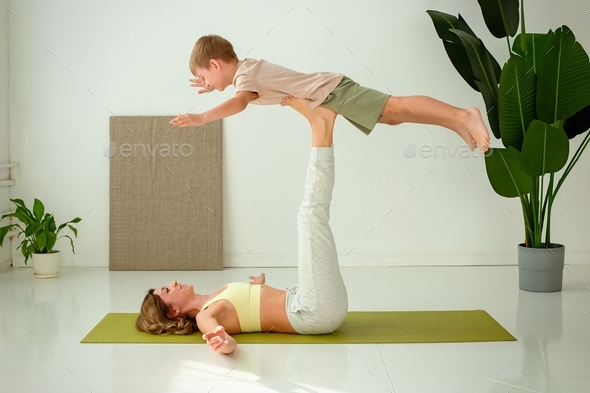 A woman, doing yoga with a child boy, lies on a sports mat, raising her legs up, doing exercises wit