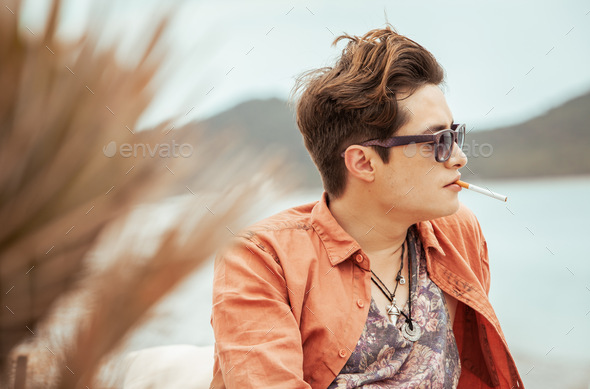 A young man dressed in an orange shirt smokes a cigarette at a lounge bar on the beach.