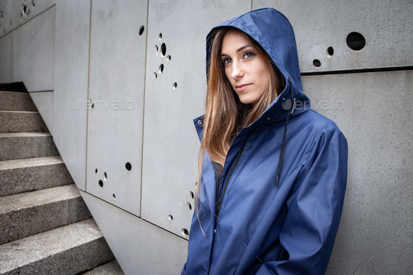 A millennial girl dressed in a blue raincoat poses thoughtfully with her hood up