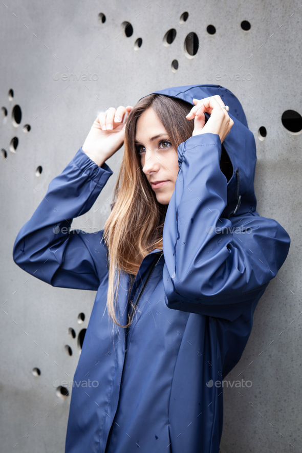 A millennial girl dressed in a blue raincoat poses thoughtfully with her hood up.