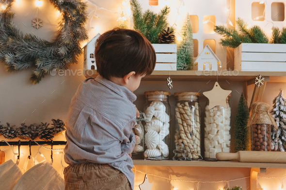 Rear view of little boy opening glass jar with sweets in kitchen decorated for christmas holidays