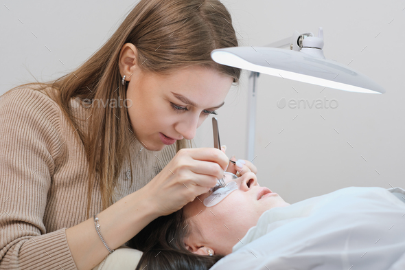 young woman beautician holding tweezers with false lash during lash extension procedure - Stock Photo - Images