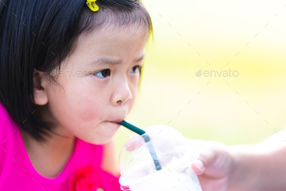Head shot of Asian kid girl sucking sweet Coconut Smoothie. Mom holding glass to feeding daughter.