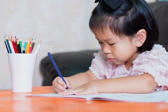 Portrait of Asian pupil girl doing homework at home. Child holding color pencil with right-hand.