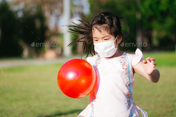 Kindergarten kid running and holding red plastic ball on public park. Child wearing medical mask.