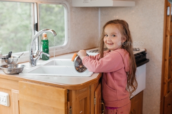 A little girl washes the dishes in the kitchen in the motorhome. Happy childhood concept