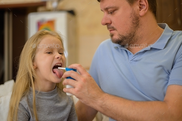 dad squirts spray medicine down the throat of his sick child on the couch at home