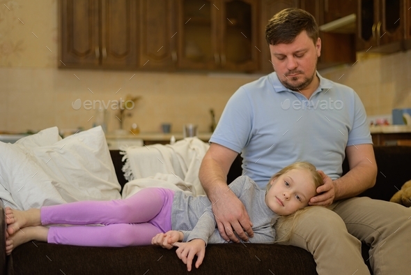 a sick child lies on the couch with his dad\'s head on his lap, who strokes her on the head