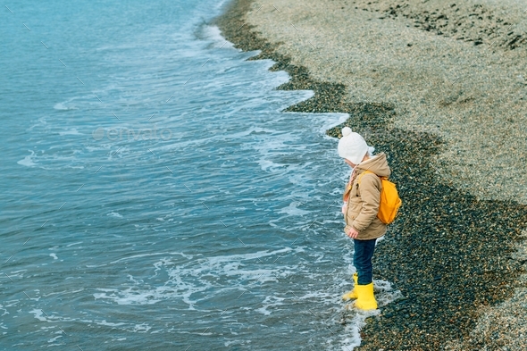 little girl stands in profile in outerwear on the rocky coast of the sea, ocean in autumn, spring