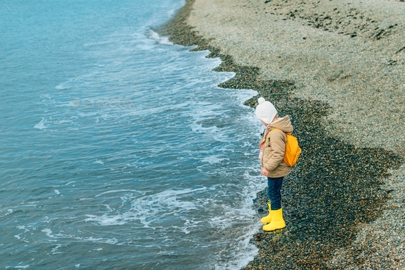 little girl stands in profile in outerwear on the rocky coast of the sea, ocean in autumn, spring