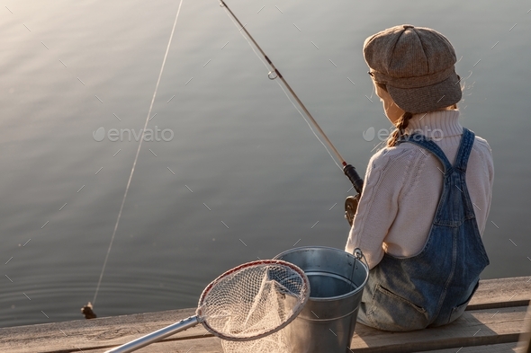 a girl in a cap with a bucket and a net is fishing on a lake with a wooden pier at sunset