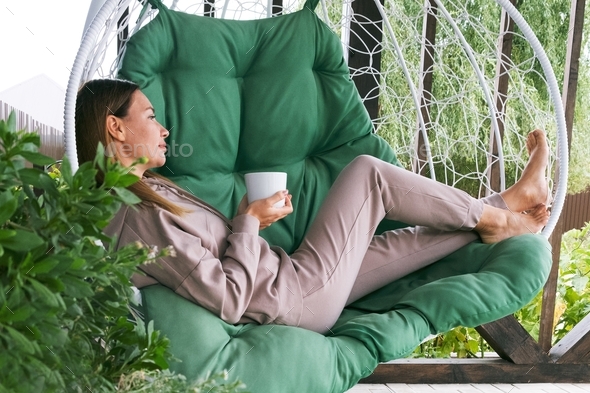 Real woman drinking tea or coffee outdoor. full length mature woman sitting in a hammock chair with