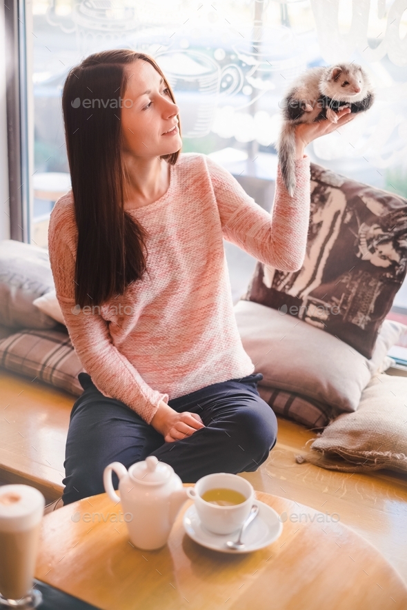 Happy young woman holding high her pet ferret in cafe