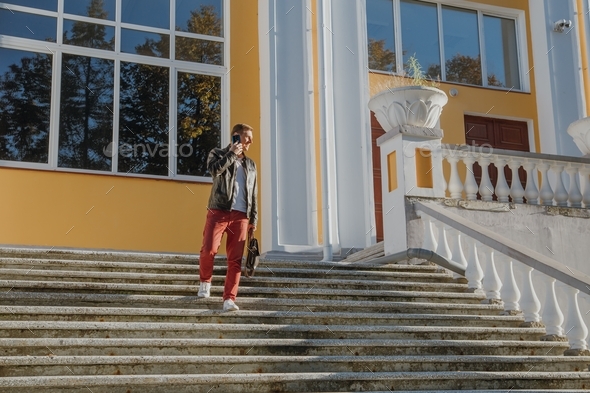 a young man walks down the stairs and talks on the phone near a classical-style building