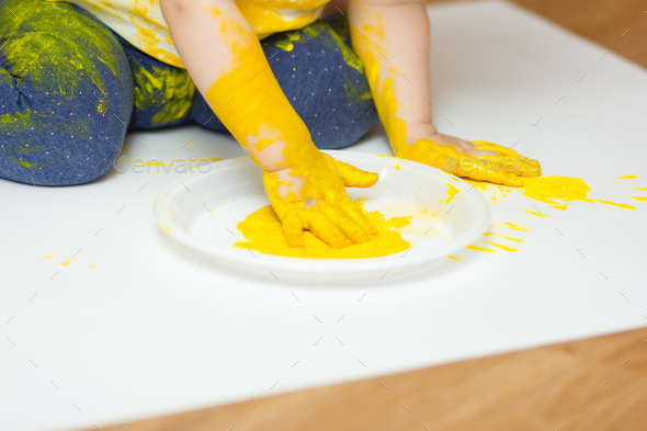 The baby dips his hand in yellow paint poured on a white plate.